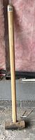 Full view of the sledgehammer showing the wooden handle with metal collar and the heavy metal head resting on a floor surface.