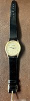 Photo of Tekron wristwatch showing round gold-tone case, cream dial with black numerals, and black leather strap with buckle.