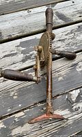 Photo of a vintage hand crank drill featuring wood handles and a shoulder plate brace resting on a weathered wooden surface, showing rust and mechanical detail.