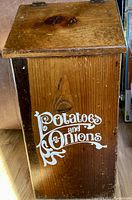 Photo of vintage wooden box with hinge lid, featuring white decorative text 'Potatoes and Onions' on the side, showing wood grain and signs of age.