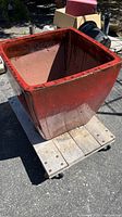 Angled front view of large square glazed red terra-cotta planter sitting on a wooden dolly, showing its size and exterior condition.