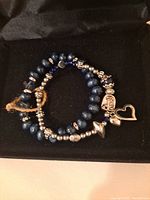 Two expandable handmade bead bracelets, one with dark blue and silver-tone beads with multiple silver tone charms including heart outline and angel figure. The second bracelet also features blue and silver-tone beads with a heart charm, resting at 2 1/2 inches diameter.