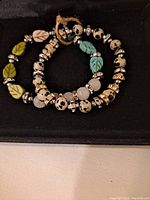 Two natural stone bracelets shown together on a black surface, showing dalmatian jasper, rose quartz, green and turquoise leaf beads, and silver spacer beads with rhinestones.