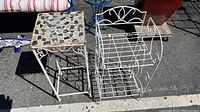 Two white metal outdoor side tables/plant stands shown outdoors on asphalt surface, one with mosaic tile top and fixed legs, one foldable with two wire grid shelves and decorative sides.