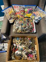 Multiple large clear resealable bags and cardboard boxes filled with assorted broken and whole jewelry and beads placed on a gray armchair.