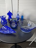 Photo showing full set of seven blue glass vases and bowls on black round table with a white shutter background.