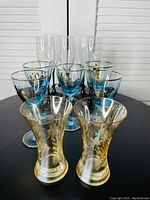 Ten decorative drinking glasses arranged on a black surface - four tall clear ribbed glasses, four blue stemmed glasses with gold and black flower motifs, and two smaller amber etched glasses.