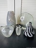 Five decorative glass items arranged together including a tall vase, a small textured vase, a heart-shaped bowl, a round platter, and a black and white striped vase.
