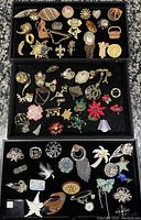 Overview of three trays filled with assorted vintage and modern brooches including varied designs, materials, and colors.