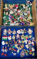 Top view of two trays filled with various Christmas-themed brooches and pins showing many different holiday related designs in a cardboard box and on a blue fabric.
