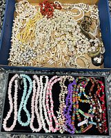 Top view of a box containing various shell necklaces and bracelets including dyed shell chips, puka shells, cowrie shells, and colorful multicolored beads.