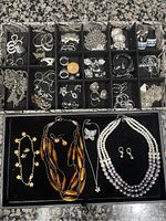 Overview of multiple trays with a variety of vintage and modern necklaces and earrings. Includes layered pearl necklace, brown beaded necklace, silver butterfly pendant, and multiple pairs of earrings.