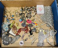 Top view of cardboard box with assorted jewelry pieces including necklaces, earrings, pins, brooches, and chains. Various colors and materials visible such as metal, beads, and stones.