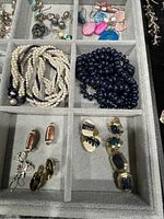 Photo showing assorted earrings and two multi-strand necklaces, one in faux pearls and one in navy blue beads, held in a gray fabric tray compartment.