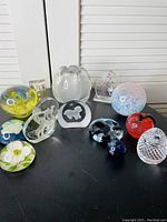 Full view of eleven assorted art glass paperweights on black surface showing shapes and colors including yellow, red, clear, and blue pieces