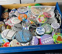Box filled with a diverse collection of pins and badges, showing a variety of designs including birthday wishes, political statements, and humorous phrases.
