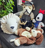 Five plush toys displayed on a couch: bulldog in overalls and cap, small black and tan dog, brown and white floppy-eared dog, white lion with fabric mane, and Snoopy in plaid shirt with antlers.
