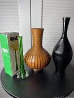 Three vases on a black surface including a tall green glass vase in original box, a woven brown vase, and a tall black vase.
