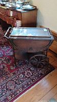 Side view of antique wood drop leaf tea cart with decorative carvings and large wooden wheels on patterned rug.