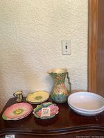 Full assortment of plates, jug, bowl and small vase displayed on dark wood furniture against textured white wall and wooden door frame