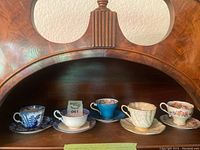 Five assorted tea cups with matching saucers arranged side by side on wooden shelf