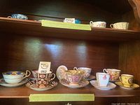 Photo showing assorted tea cups and saucers arranged on a wooden shelf, various designs and colors visible.