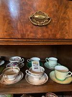 Photo of shelf with six assorted vintage ceramic teacups and saucers arranged in two rows, showing floral and patterned designs with gold rims.