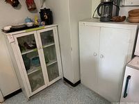 Two white cabinets side by side in a room with visible wear and chips. Left cabinet has glass doors showing contents inside, right cabinet has solid doors.