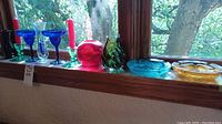 Window ledge displaying assorted colored glass items including cobalt blue glass stemware, clear glass vase, red round glass decorative piece, green swirled vase, and pairs of green glass candle holders with red candles.