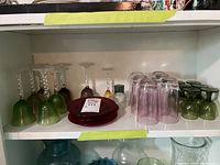 Shelf display showing multiple coloured glasses and plates: green glasses upside down in the front, purple glasses with twisted stems, red plates in the center, pinkish inverted glasses on the right.