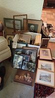 Wide view of assorted framed photos and art leaning against wall and furniture, including various sizes and frame styles.