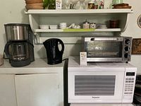 Overview of coffee maker, electric kettle, toaster oven, and microwave on counter beneath shelf with kitchen items