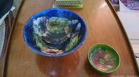 Image of both Moorcroft pottery dishes on a wooden surface showing floral patterns; larger blue bowl and smaller green dish.