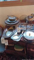 Photo of assortment of silver plated serving items on wooden table including trays, bowls, teapot, covered dishes, bread basket, and strainer.