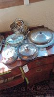 Photo showing assortment of silver plated serving items on a wooden surface including covered dishes, pedestal dish, urn warmer