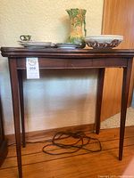 Front view of the wooden folding top table showing the four legs and apron with grooved detailing. Several decorative ceramic items are on the tabletop.