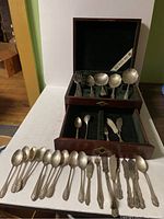 Silverware displayed partially in two-tier wooden box with green fabric lining, resting on a white table with pieces laid out in front.
