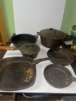 Six cast iron pans of varying sizes including skillets, grill pan, covered pan, and pot with lid, displayed on a surface against a white backdrop.