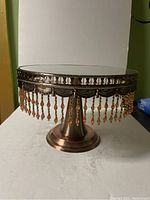 Copper-colored metal pedestal stand with mirrored top and hanging amber-colored beaded fringe around the edge, photographed from front view.