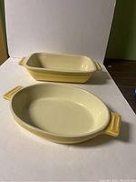 Two cream-colored ceramic oven dishes, one rectangular and one oval, both with yellow handles, shown from an angle.