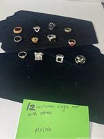12 costume rings displayed on a black background, each ring varies in stone color, shape, and metal finish.