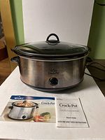 Front view of Rival Crock-Pot with stainless steel body, black trim, glass lid, control dial, and user manuals placed in front.