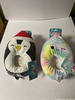 Two kids' size 11/12 Squishmallows slippers, one penguin wearing a Santa hat and one pastel rainbow tie-dye character, both new with tags.