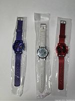 Three wristwatches in sealed plastic sleeves: one blue, one white, and one red, laid side by side on a white surface.
