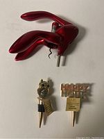 Red metallic wing-shaped wine bottle opener with corkscrew shown from top angle, along with owl and 'Happy Hour' wine stoppers