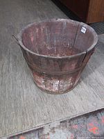 Photo showing angled view of old wooden sap bucket on wooden floor, highlighting its round shape, metal handle, and aged wood surface.