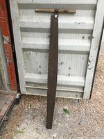 Full view of the antique one-handled metal saw standing vertically against a container door, showing the full length and condition of the blade and handle.