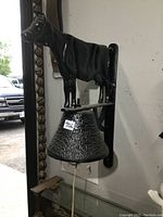 Front and side profile of the vintage cast metal cow bell mounted on the wall, showing the detailed cow figure above the textured bell and the pull rope hanging down.