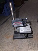 Front-side view of Dremel Model 210 drill press showing upright column, adjustable table, and drill clamp with white levers.