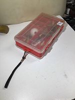 Red double-sided fishing tackle box with transparent lid showing multiple compartments filled with various fishing lures, jigs, and tackle items.